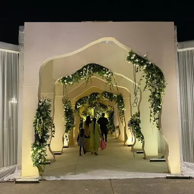 A modern and artistic entrance structure for an evening reception. The custom-built, free-flowing arched tunnel is accented with delicate white florals and uplighting, creating a sculptural and impressive welcome.