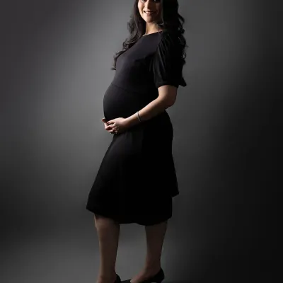 A full-length silhouette shot that beautifully defines the baby bump. The mom-to-be stands in a simple black dress against a grey backdrop, creating a timeless and elegant maternity portrait.