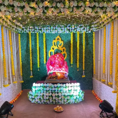 This is a larger traditional setup featuring a full enclosure with an artificial grass wall and a ceiling covered in white flowers and marigold garlands. The design creates a complete mandap structure, giving the feeling of a temple at home.