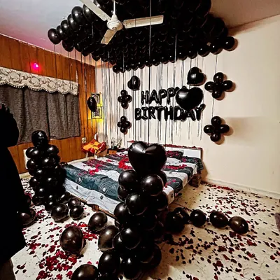 A dramatic and romantic all-black themed room decoration. This setup includes black balloons covering the ceiling, heart-shaped foil balloons, and rose petals on the floor and bed.