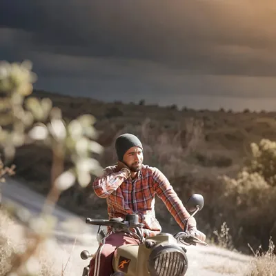 An old-school, rugged portrait of a man on his motorcycle. The dramatic, cloudy sky and earthy tones give this photo a cinematic and adventurous feel.