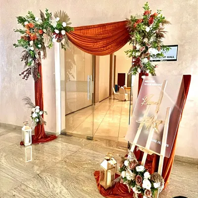 A grand entrance for a wedding hall, styled with an asymmetrical arch of rich fabric and florals, lanterns, and a custom acrylic welcome sign.