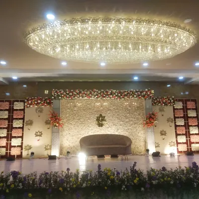 This is a wide view of a reception stage I designed, featuring a massive crystal chandelier overhead. The backdrop is a full wall of white flowers with a central Ganesha motif and lotus accents.