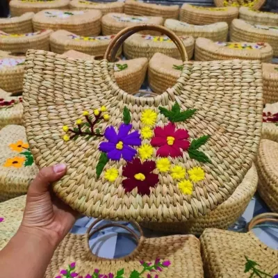 A closer look at one of our fan-shaped sabai grass bags, featuring colorful and detailed floral embroidery. Held in hand, you can see the fine craftsmanship and sturdy wooden handle that make this artisan bag both beautiful and practical.