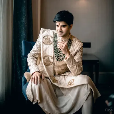 A classic, regal portrait of the groom seated, looking poised and ready for his wedding. The lighting highlights the rich texture of his traditional sherwani.