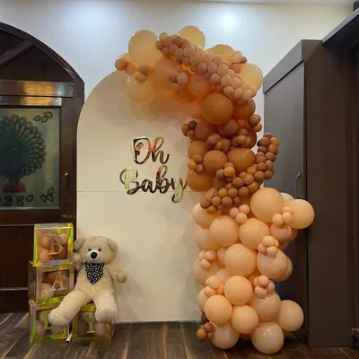 A full view of our 'Oh Baby' shower decoration. The setup includes a white arched backdrop with gold lettering, a cascading balloon arch in neutral tones, a teddy bear prop, and transparent 'BABY' letter blocks.