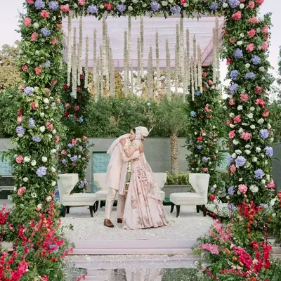 A beautiful moment of embrace under a lush floral mandap. The structure is covered in pink, purple, and red flowers, with hanging tuberoses adding a traditional touch to the modern pastel theme.