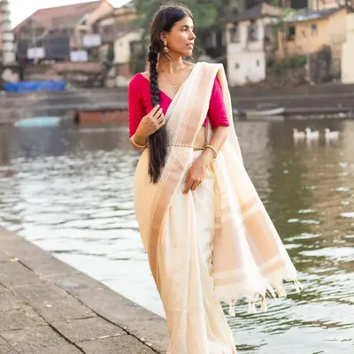 A poised, full-length shot of a classic off-white and pink silk saree, showcasing its elegant drape and texture against a scenic riverside backdrop.