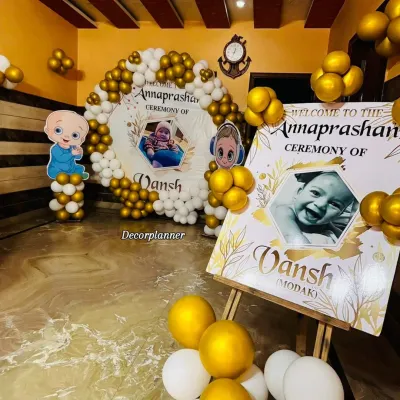 A complete view of the personalized Annaprashan ceremony for baby Vansh. The setup includes a custom welcome easel and a matching circular backdrop, both featuring his photo, framed by a beautiful white and gold balloon arch.