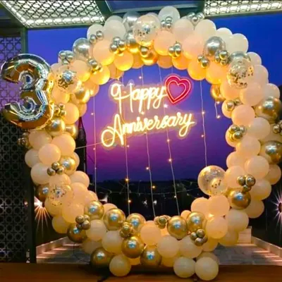 A beautiful anniversary decoration featuring a balloon ring in white, gold, and transparent confetti balloons. The 'Happy Anniversary' neon sign and fairy lights create a warm and celebratory glow.