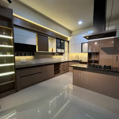 This island kitchen showcases a beautiful combination of high-gloss acrylic surfaces and a duo-tone color scheme. The lit-up glass display cabinet and sleek black chimney add to the modern and balanced output of this design.