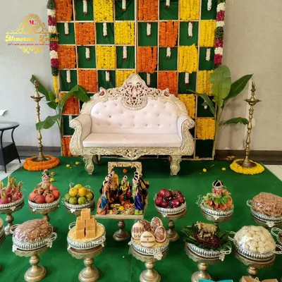 A traditional setup for a baby shower or naming ceremony, featuring a backdrop of marigold flowers and banana leaves, an ornate sofa, and brass lamps (diyas).