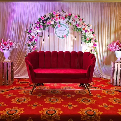 A stunning stage setup for an anniversary or engagement party. A plush, shell-shaped red velvet sofa sits before a beautiful semi-arch of pink and white roses, set against a backdrop of elegant white curtains.