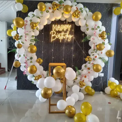 An elegant and classic birthday backdrop featuring a full circle balloon garland in white and gold. The setup is enhanced with a 'Happy Birthday' neon sign and a small pedestal for the cake.
