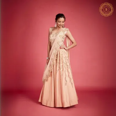 Own the moment in this elegant peach Indo-western gown. The attached drape gives it the grace of a saree with the comfort of a gown.