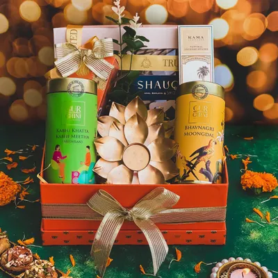 The "Raunaq" hamper is designed to bring splendor to any Diwali celebration. This vibrant orange box includes gourmet Indian snacks, natural incense sticks, and a stunning gold-plated lotus diya as the centerpiece.