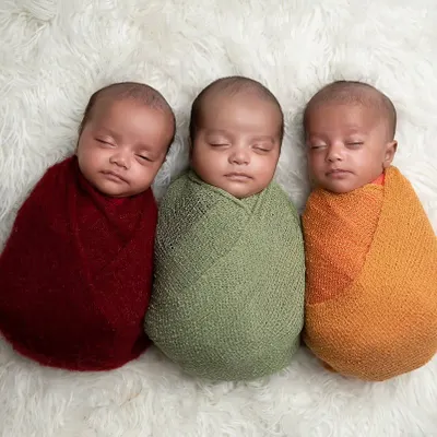 Three times the love. Here are beautiful newborn triplets, each swaddled in a different warm color, sleeping peacefully side-by-side.