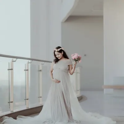 A full-length portrait of an expecting mother in a stunning, light-colored maternity gown, holding a bouquet of pink roses. This elegant maternity photo was taken in a bright, modern indoor location.