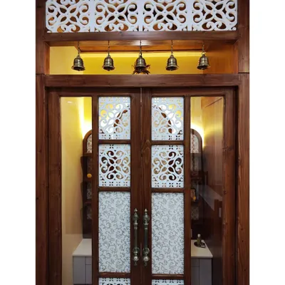 The doors to a divine space. This pooja room entrance features custom wooden doors with frosted glass, intricate jaali cutouts, and traditional brass handles. The transom window above with hanging bells adds a final touch of temple-inspired elegance.