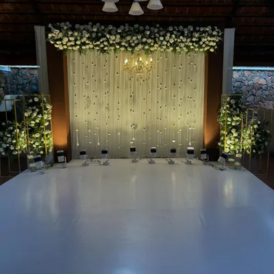 This cozy and elegant setup is perfect for an engagement or reception. The backdrop features a white drape with hanging jasmine buds, topped with a dense arrangement of white roses and a chandelier.