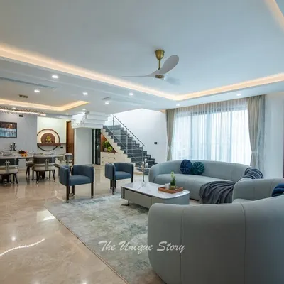 An open-plan living and dining area designed for a modern contemporary home in Jakkur. The space features soft, curved sofas, a sleek marble coffee table, and an elegant 8-seater dining set, all unified by polished marble flooring and sophisticated cove lighting.