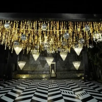 A breathtaking Sangeet stage, dripping with glitz and glam. The ceiling is a cascade of golden tassels, disco balls, and crystal chandeliers over a bold black-and-white checkered dance floor.