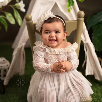 A beautiful sitter portrait of a baby girl with a heartwarming smile. Dressed in an elegant white lace outfit, she sits confidently in front of a charming tent backdrop, perfectly capturing this milestone.