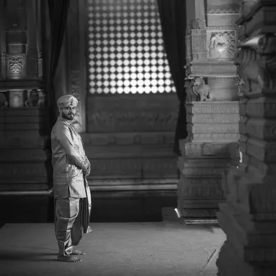 A groom's portrait in black and white, set against the timeless stone pillars of a temple. The dramatic lighting and his solitary figure convey a sense of quiet strength and contemplation.