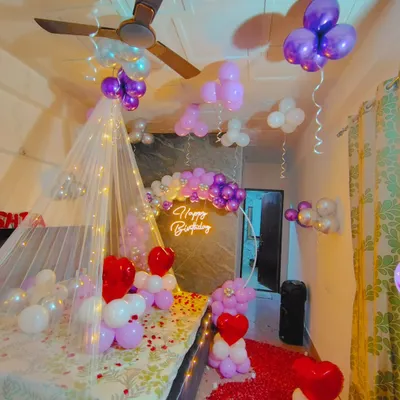 A romantic birthday room setup with a fairy light canopy, a rose petal path, and a purple and white balloon backdrop with a neon sign.