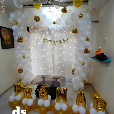 An elegant white and gold balloon arch against a fairy light curtain, personalized with the name 'PRANAB' in foil balloons at the base. This is a classic choice for a sophisticated home party.