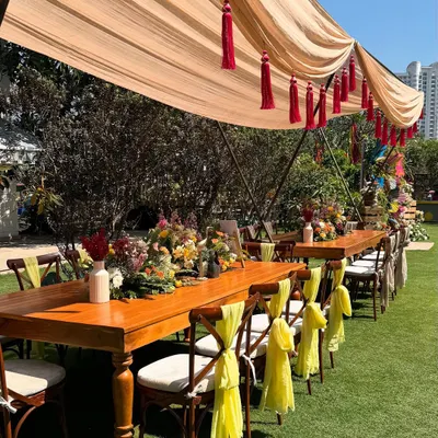 A beautiful long table set for an outdoor Holi lunch. The wooden table is decorated with a runner of fresh, colorful flowers, and the chairs are adorned with bright yellow fabric ties, adding to the festive atmosphere.