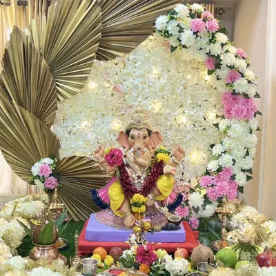 A stunning Ganpati decoration featuring a backdrop of white flowers and fairy lights, framed by large golden palm leaves for a modern, divine look.