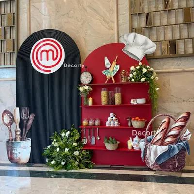 I create bold, thematic photo opportunities like this one for my Masterchef events. The setup includes a custom red shelving unit with cooking ingredients, a branded backdrop, and fun props like a chef's hat.