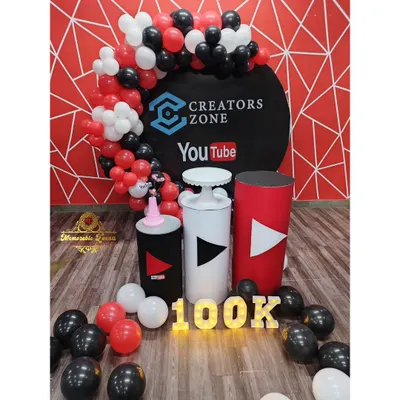 This is the full setup for the Creators Zone 100k subscriber celebration. It includes a branded backdrop, YouTube-themed pedestals, and marquee lights, all framed by a red, black, and white balloon garland.
