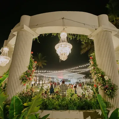 A grand entrance arch designed with stately white pillars, adorned with floral arrangements and magnificent hanging chandeliers. This entrance opens up to an outdoor reception, promising a night of elegance and celebration.
