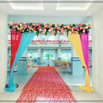 A bright and cheerful entranceway for a wedding hall, decorated with colorful drapes in red, blue, yellow, and pink. A lush floral arrangement at the top completes this festive welcome for your guests.