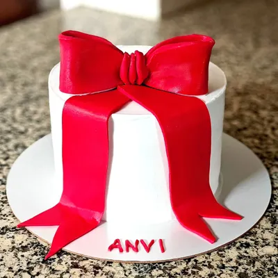 The core of elegance is simplicity. This cake features a clean white buttercream finish and a striking, handcrafted red fondant bow, personalized with the name 'Anvi'.