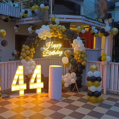 A stylish outdoor terrace decoration for a 44th birthday. I used a black, yellow, and white balloon arch, a 'Happy Birthday' neon sign, and large marquee numbers to create a festive photo corner.