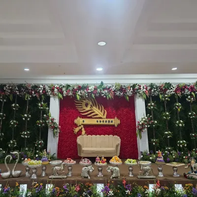 A unique stage design with a Krishna theme, featuring a large golden flute and peacock feather against a wall of red roses.