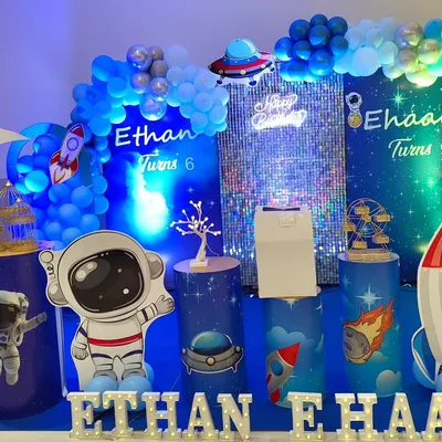 A futuristic space theme with cool blue lighting and a shimmering sequin wall. The setup is filled with astronaut and rocket cutouts for a cosmic feel.