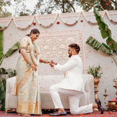 A beautiful proposal moment during a traditional engagement ceremony. The backdrop is a pristine white panel with intricate carvings, flanked by banana plants and traditional brass lamps, creating a purely elegant and culturally rich setting.