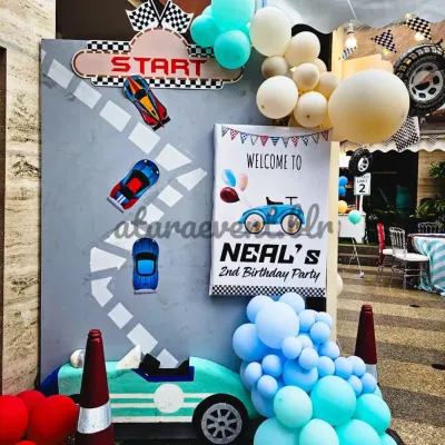 The race begins here! A "Welcome to Neal's 2nd Birthday Party" sign sets the stage, complete with a custom racetrack backdrop, car cutouts, and a blue balloon garland.