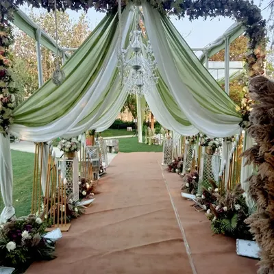 A breathtaking entrance for a day wedding, with a walkway covered in green and white drapes, floral arrangements, and elegant chandeliers.