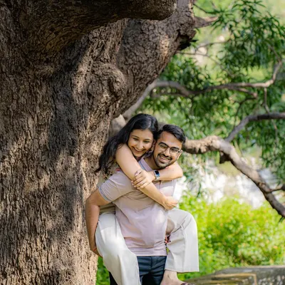 A playful and happy moment with a piggyback ride under a large, beautiful tree. This candid shot from an outdoor pre-wedding session is full of natural joy and laughter.