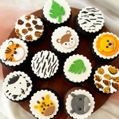 A dozen jungle safari cupcakes on rich chocolate bases. This set features a mix of fondant animal faces like a tiger, giraffe, and zebra, alongside animal print designs and monstera leaves for a complete jungle party theme.
