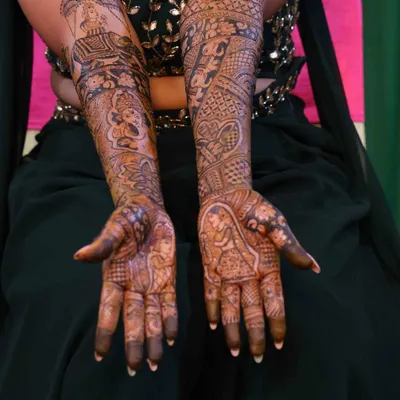 A beautiful example of a full bridal mehndi after the stain has set. The design covers both arms with a rich, dark color, featuring a mix of portraits, traditional patterns, and personalized story elements.
