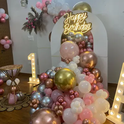 A beautiful and modern first birthday setup with a boho-chic feel. This decoration uses a cascade of rose gold, gold, silver, and white balloons flowing from an arched backdrop, complemented by a neon sign and marquee initials.
