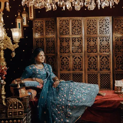 A regal solo portrait of the bride-to-be in a stunning blue lehenga, seated on an ornate set with rich, warm lighting.