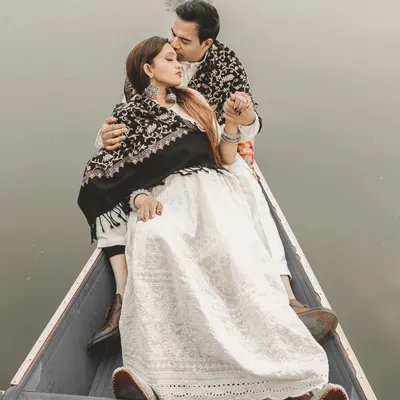 A close-up, tender moment on a Shikara boat in Kashmir. The groom-to-be wraps his partner in a traditional shawl, creating a warm and intimate portrait against the cool, misty backdrop of Dal Lake.