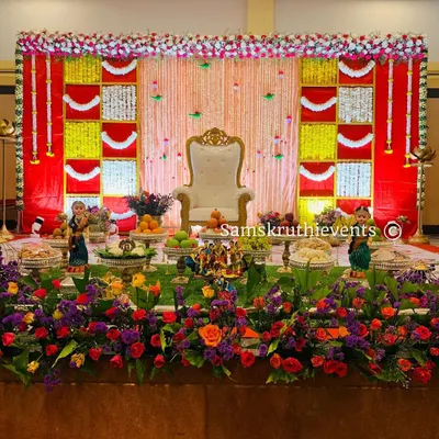 A traditional Seemantha decoration with a vibrant red and yellow checkered backdrop. The stage is beautifully framed with a thick border of fresh flowers.
